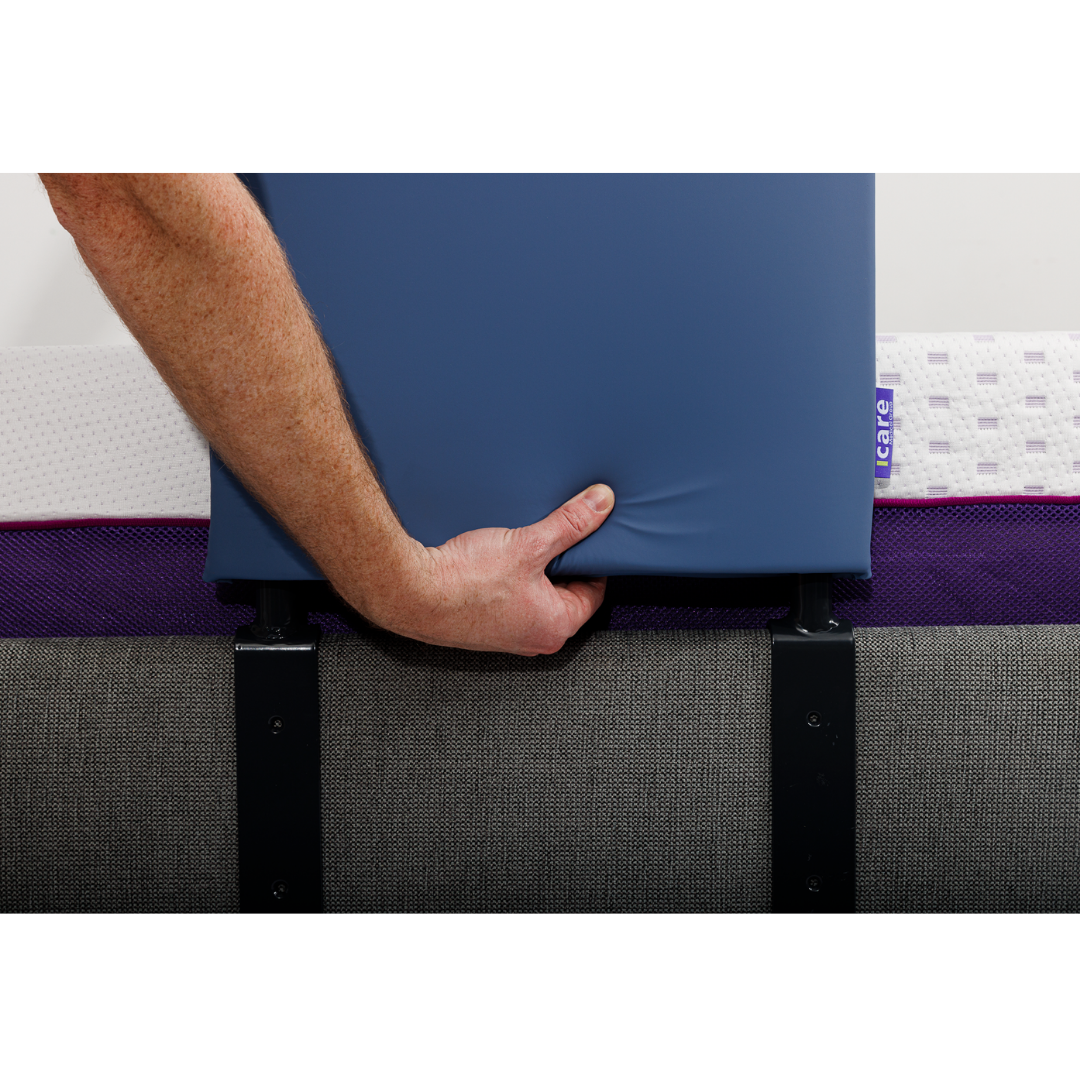A person’s hand holds the iCare Medical Bed Safety Rail, a sturdy blue support attached to a bed with a gray upholstered base and white mattress for fall prevention.
