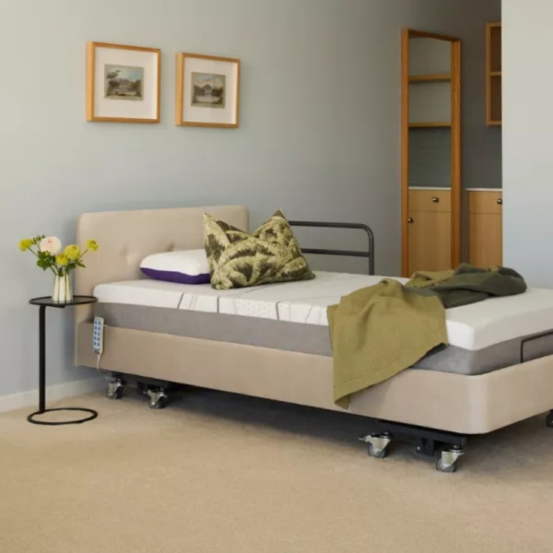 A modern bedroom with a beige adjustable bed on wheels featuring iCare Medical Bed Safety Rails, green and patterned pillows, a green blanket, a black side table with flowers, and two framed pictures on a light gray wall.