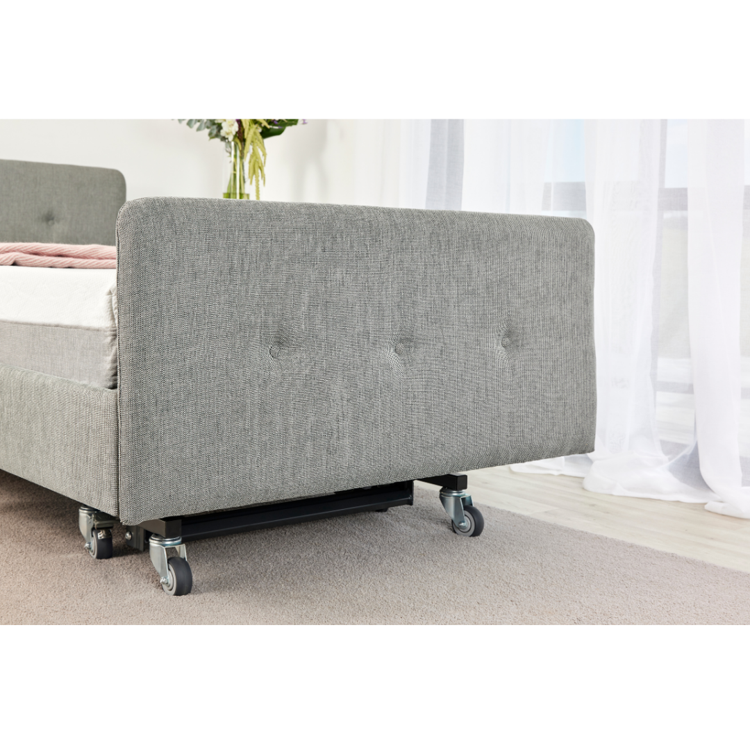 A close-up of the iCare Medical Headboard and Footboards for iCare Beds, shown in grey upholstery with wheels on a light carpet, near a window with sheer curtains and a vase of flowers in the background.