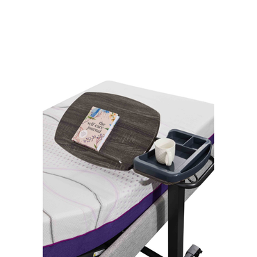 An iCare Medical Over Bed Tilting Table—height adjustable—holds a book, mug, and tray on an adjustable bed with white and purple bedding, creating a convenient multi-purpose surface.