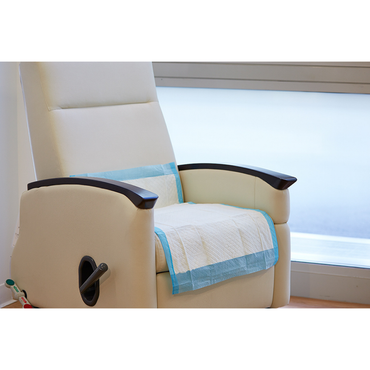 A beige medical recliner with wooden armrests is covered by a Medline Protection Plus Disposable Underpad, featuring blue and white colors and polypropylene backing for moisture protection, positioned by a frosted window in a healthcare setting.