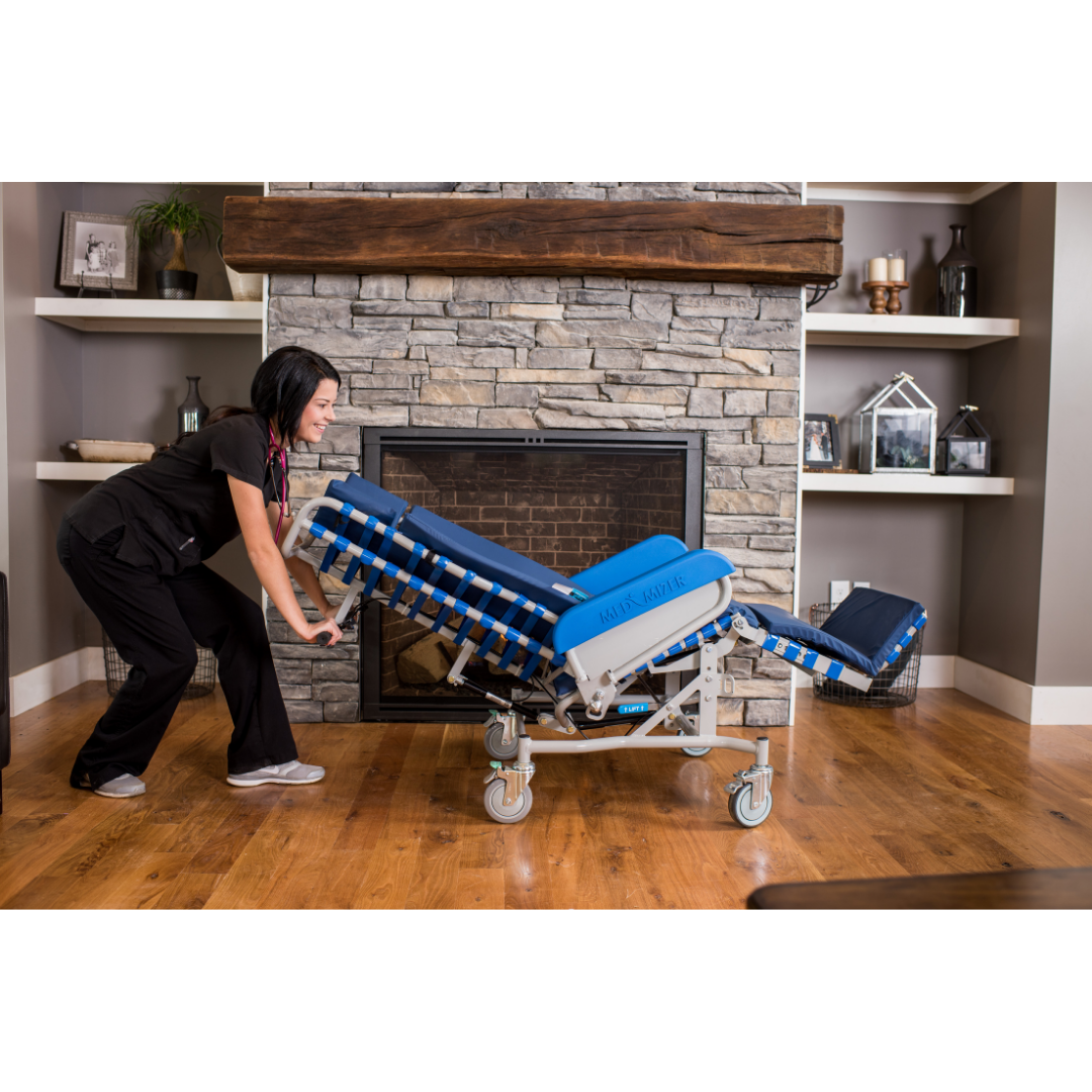 A woman in black scrubs adjusts a blue and white Med-Mizer FLEXTILT Reclining Transfer Chair with wheels in front of a stone fireplace in a living room decorated with shelves and decor.
