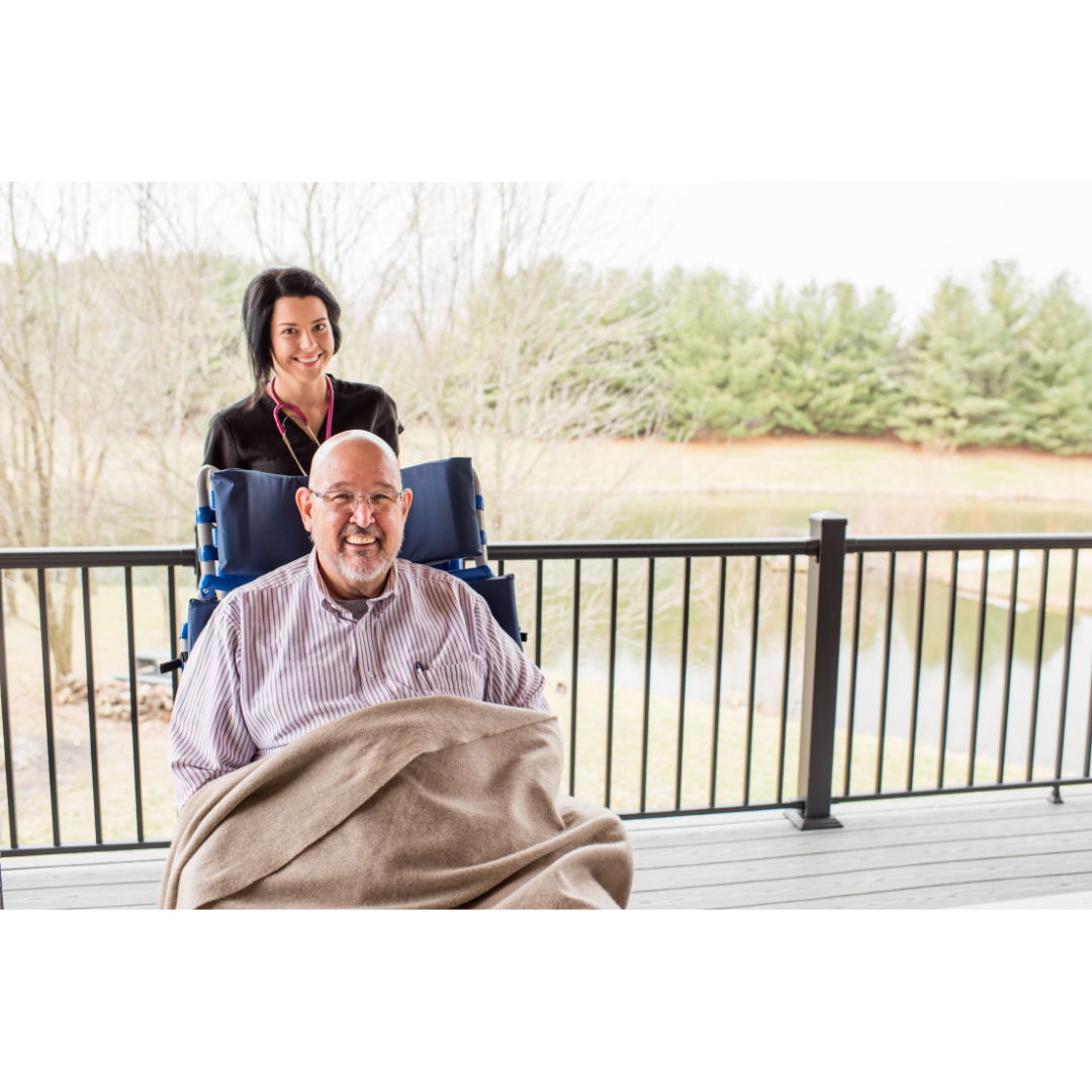 An older man smiles in a Med-Mizer FreedomFlex Reclining Transfer Chair, covered with a blanket, while a caregiver stands behind him on a porch overlooking a pond and trees on a cloudy day.