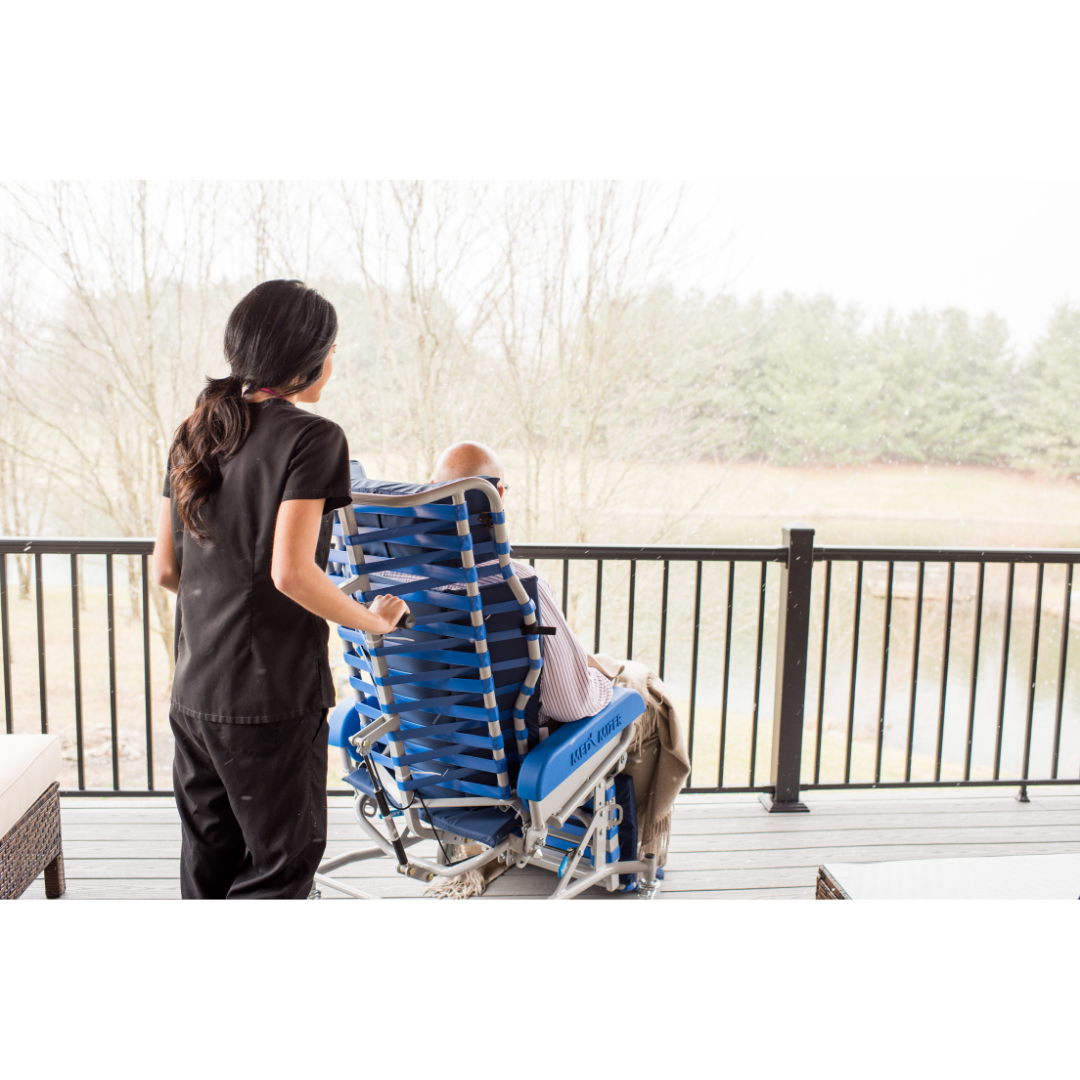 A caregiver stands behind an elderly person seated in a Med-Mizer FreedomFlex Reclining Transfer Chair on a porch, overlooking a scenic view with trees and a pond.