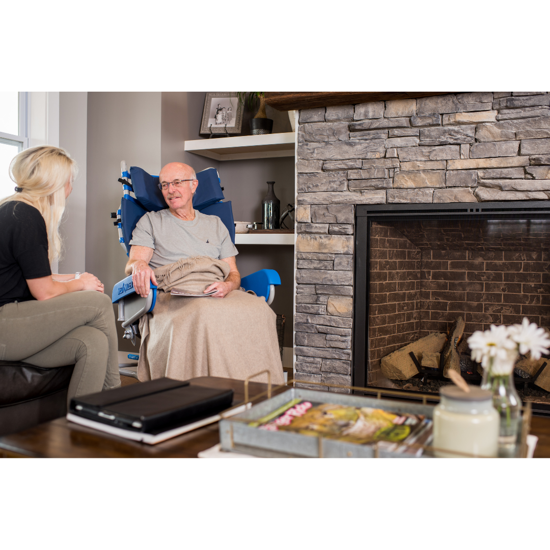 A man relaxes in a Med-Mizer FreedomFlex Reclining Transfer Chair with mobility support, smiling and chatting with a woman in a cozy living room. A stone fireplace adds warmth, and magazines and flowers sit on the coffee table.