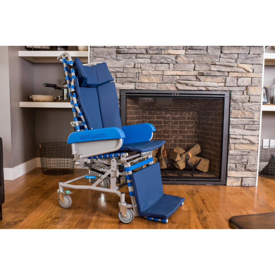 A Med-Mizer FLEXTILT Reclining Transfer Chair, a blue mobility support chair with wheels, is placed in front of a stone fireplace in a living room featuring wood floors and shelves.