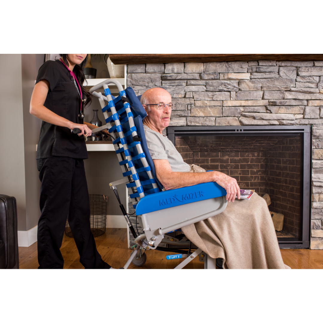An older man sits in a Med-Mizer FreedomFlex Reclining Transfer Chair with a blanket, while a caregiver in black scrubs stands behind him. They are indoors, next to a stone fireplace on a wooden floor, showing a cozy setting for mobility support.