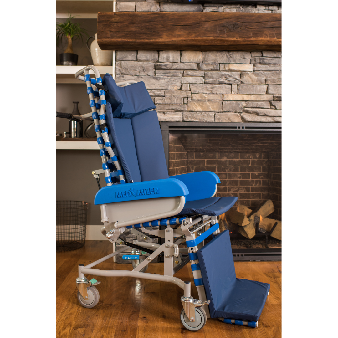 The Med-Mizer FLEXTILT Reclining Transfer Chair in blue and silver, featuring a footrest and wheels, is placed on a wooden floor before a stone fireplace in a cozy living room.
