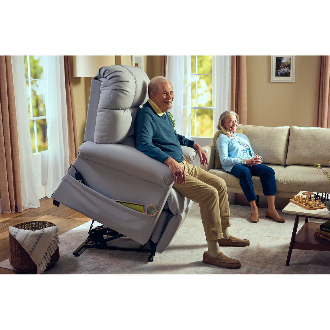 An elderly man in a blue sweater enjoys therapeutic heat and massage on his Journey Perfect Sleep Reclining Lift Chair, while an elderly woman sits nearby on the sofa. The warmly lit room with minimal decor, curtains, and a chessboard radiates comfort.