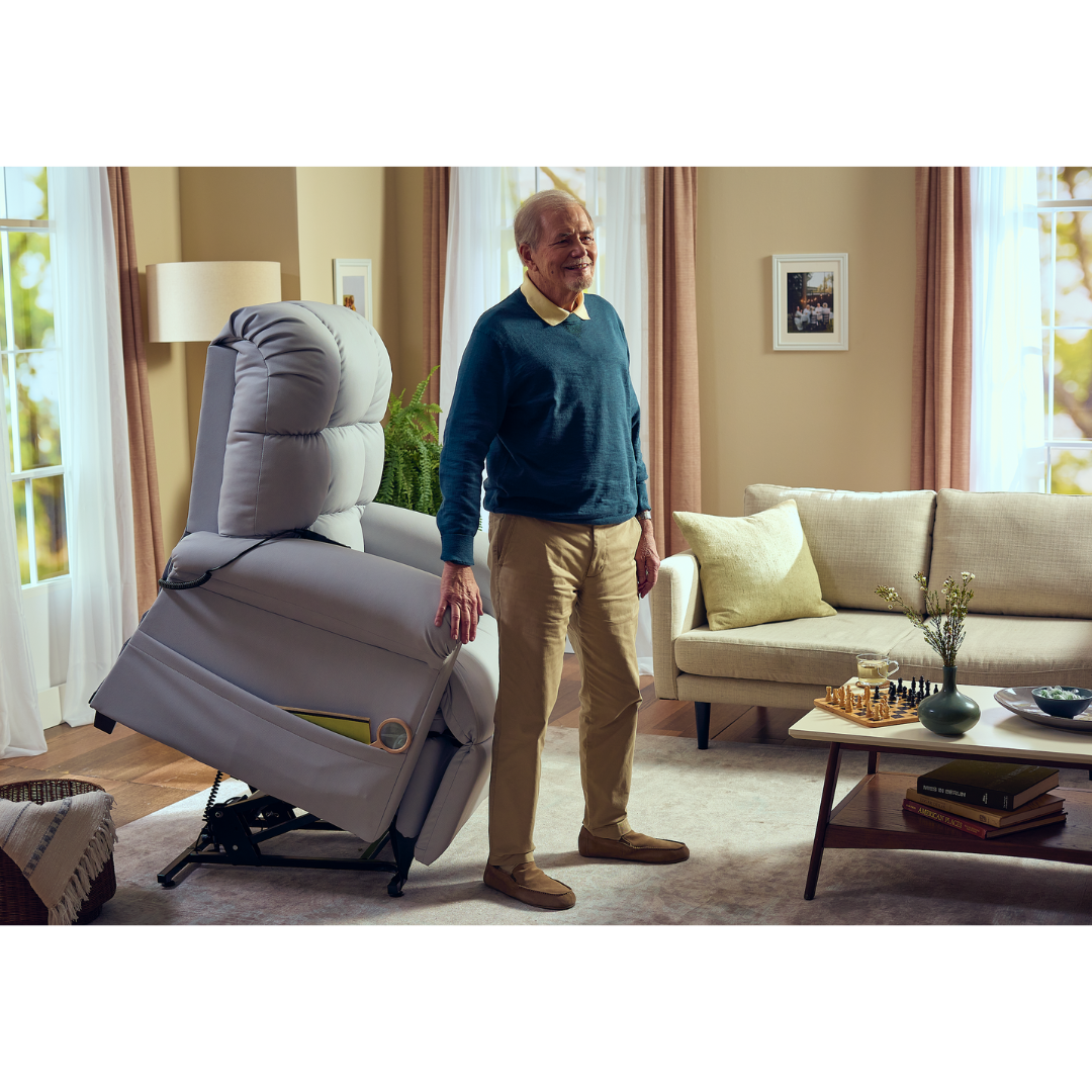 An elderly man stands next to a Journey Perfect Sleep Reclining Lift Chair with Heat in a cozy living room. The chair, partially lifted, offers therapeutic heat and massage. The space has a beige couch, decor on the coffee table, and curtains on the windows. He wears a blue sweater and khaki pants.