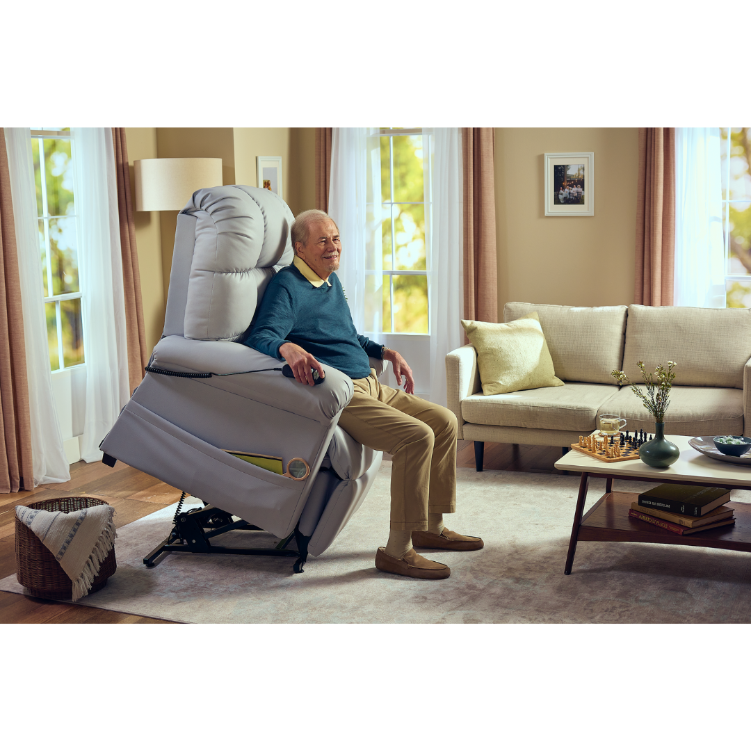 An elderly man relaxes in the Journey Perfect Sleep Reclining Lift Chair with Heat, partially elevated for comfort. The room is inviting with a cozy sofa, coffee table, and gentle lighting. Large windows showcase trees outside, amplifying the welcoming ambiance.