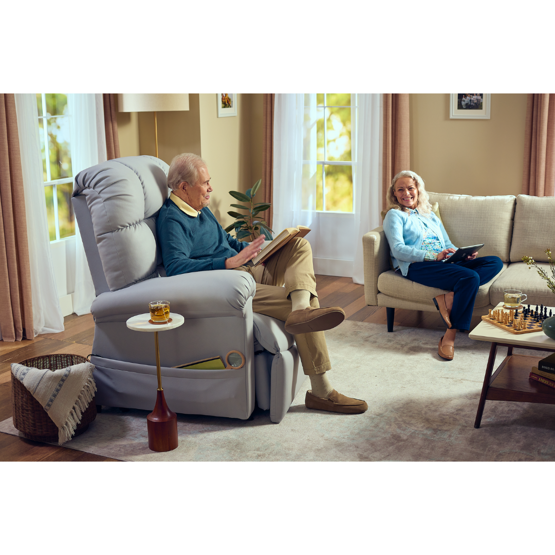 An elderly man relaxes in his Journey Perfect Sleep Reclining Lift Chair with Heat, enjoying ultimate comfort with therapeutic features, while an elderly woman smiles from the couch. The cozy living room, bathed in soft light from large windows, showcases a chess set on the coffee table.