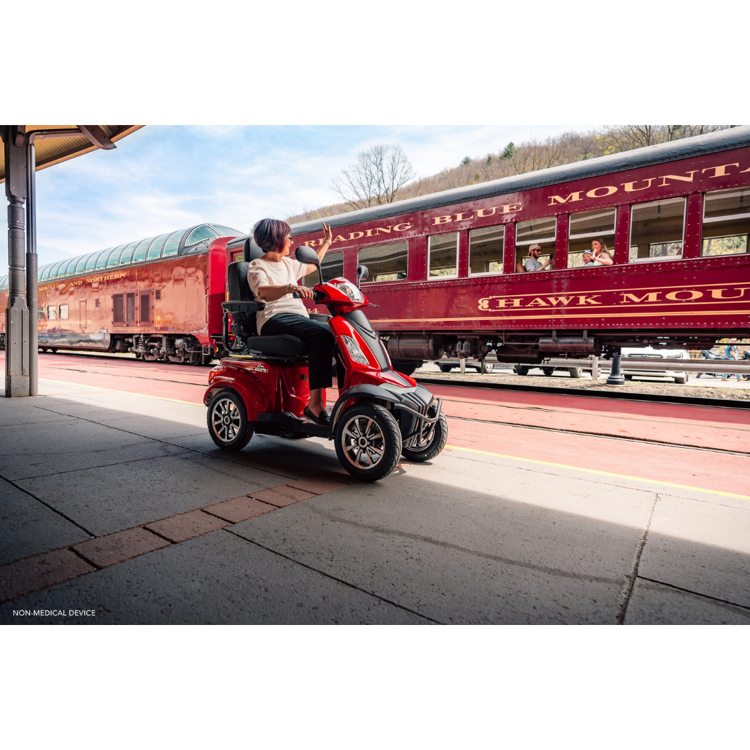 A person rides a red Pride Baja® Raptor® 2 Bariatric 4-Wheel Scooter on a sunny train station platform while passengers in a vintage maroon train look out the windows.