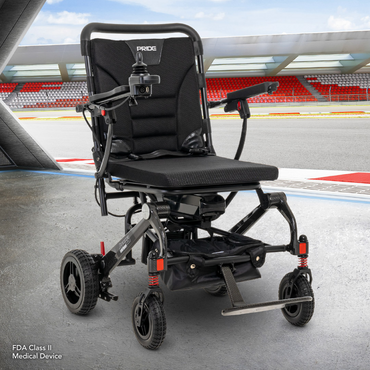 A Pride Mobility Jazzy® Carbon Ultralite Powerchair sits on an outdoor platform overlooking a racetrack. The black with red accents design features joystick control and padded seating. In the background, a stadium with red seats completes the impressive view.
