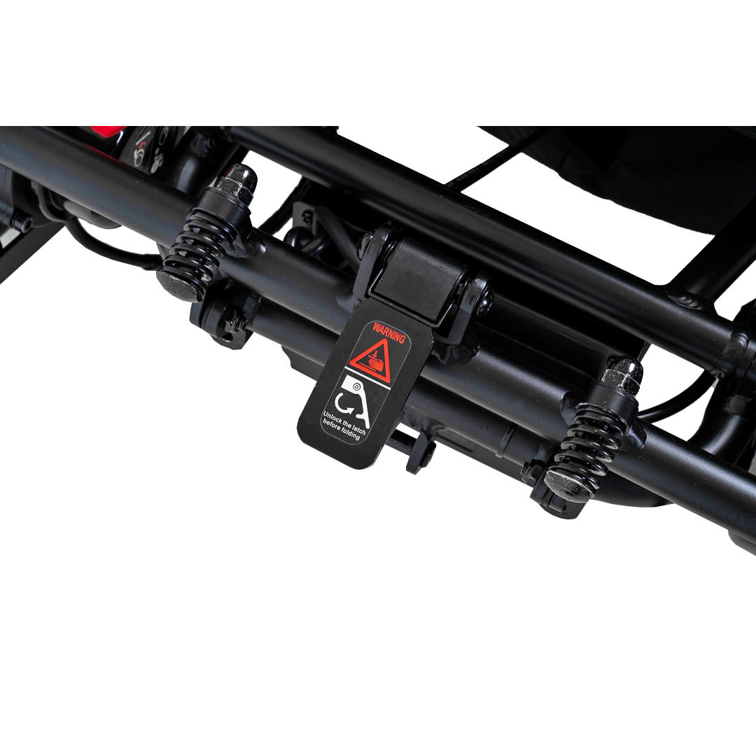 Close-up of a black mechanical suspension system with springs and a central hinge, part of the ultra-lightweight Pride Mobility Jazzy® Carbon Ultralite Powerchair. A caution label features a triangular warning symbol and crossed-out stroller image, indicating usage restrictions.