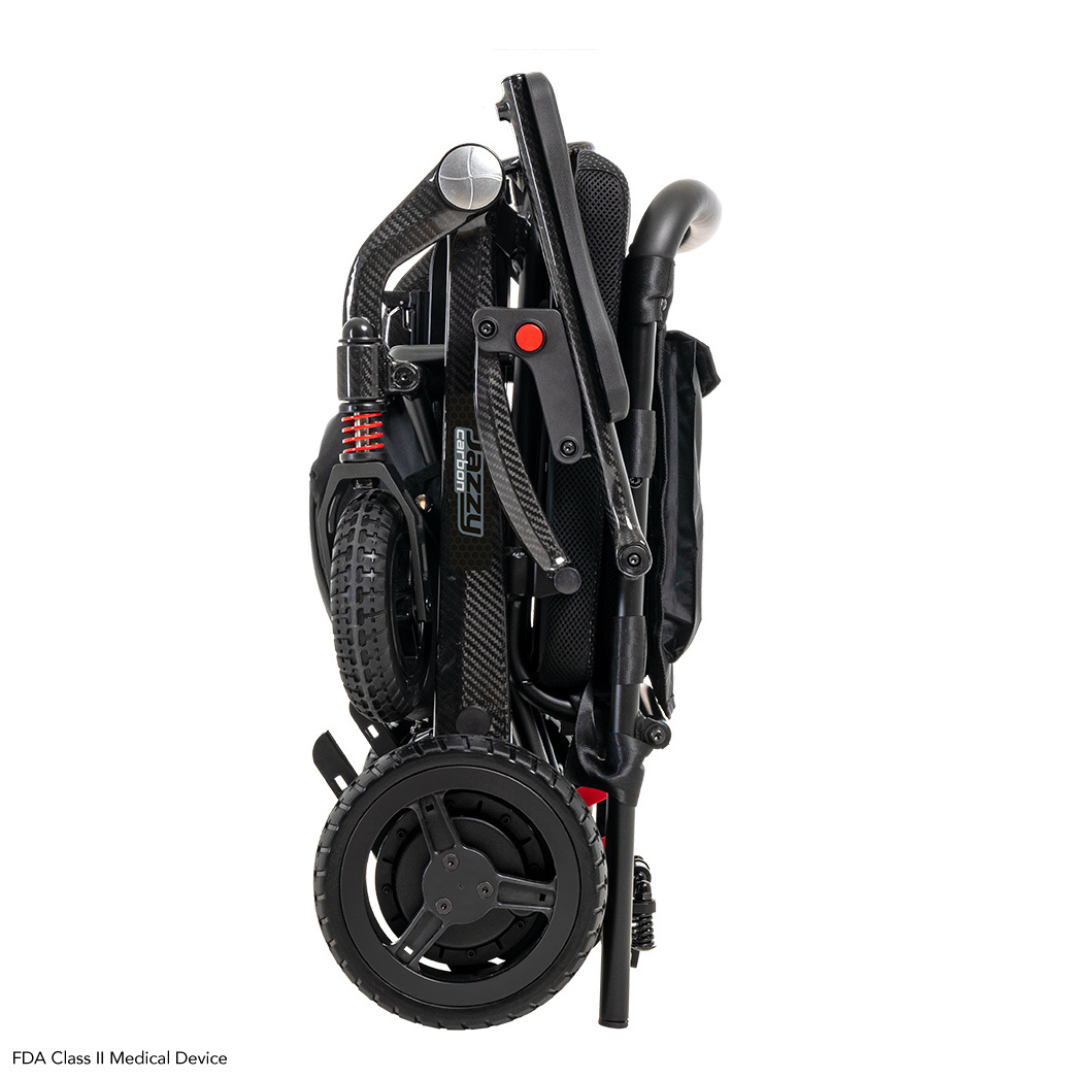 The Pride Mobility Jazzy® Carbon Ultralite Powerchair features a compact black design with large wheels and red suspension springs. It includes handles, footrests, a padded seat, and a removable joystick with USB charger. An airline-compliant lithium battery ensures convenient transport.