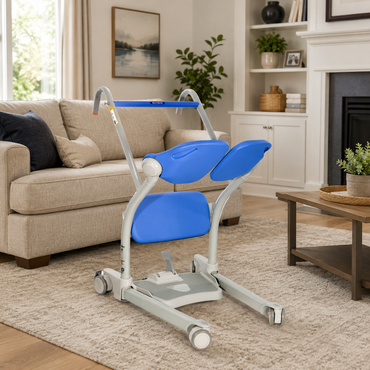 An ARJO Sara Stedy Manual Standing Aid & Sit-To-Stand Transfer Lift with blue padded supports and wheels is placed in a living room featuring a beige sofa, coffee table, and fireplace in the background.