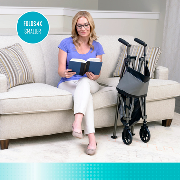 A woman sits on a sofa reading beside a folded Signature Life Elite Bariatric Travel Walker. A turquoise label states Folds 4x Smaller. The bright, homey scene features striped pillows and a soft, patterned floor.
