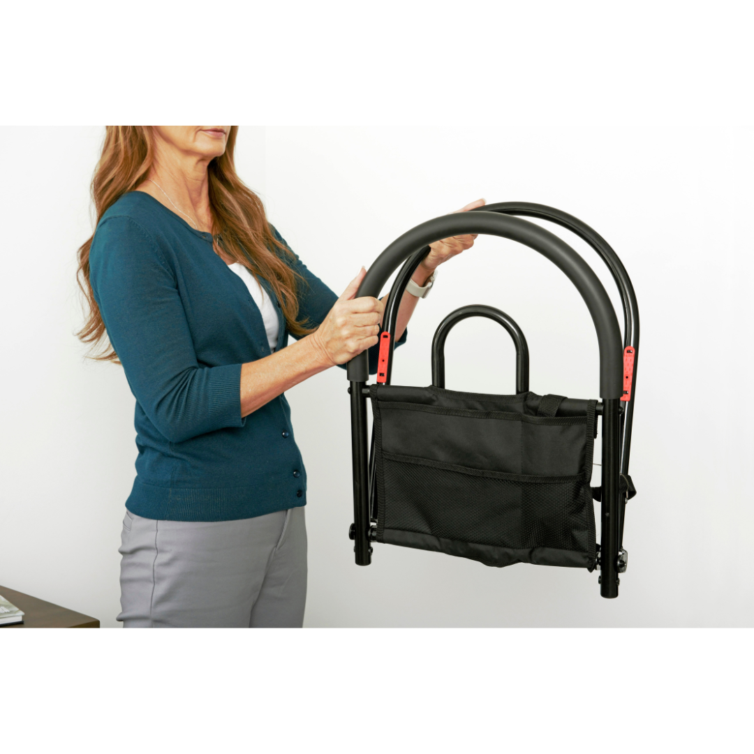 A woman in a teal cardigan holds the Stander Bed Rail Advantage Traveler, a foldable black metal bed safety rail with storage pouch—ideal for added convenience and support, shown indoors against a white wall.