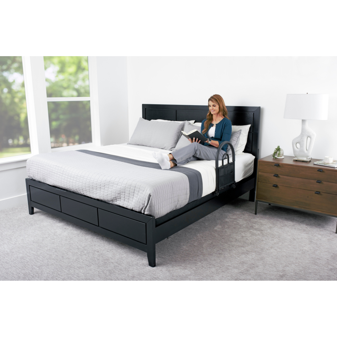A woman reads on a neatly made bed with gray and white bedding, next to the Stander Bed Rail Advantage Traveler - Portable Bed Safety Rail attached by the bedside and a wooden nightstand with a white lamp in this bright, modern bedroom.