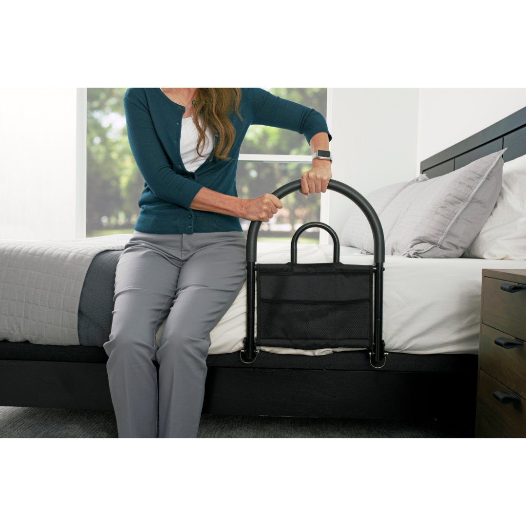 A woman sits on the edge of a bed, gripping the Stander Bed Rail Advantage Traveler - Portable Bed Safety Rail attached to the mattress in a bright bedroom.