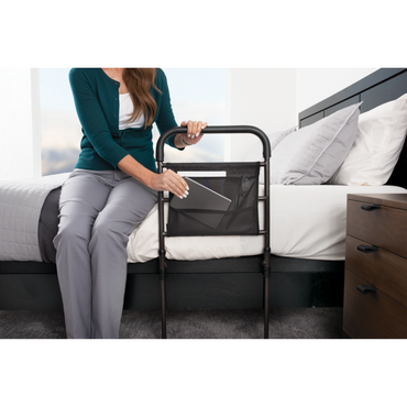 A woman sits on the edge of a bed, placing a book into the pocket of the Stander Stable Rail - Bedside Standing Aid with Extendable Legs, positioned next to her in a modern bedroom.