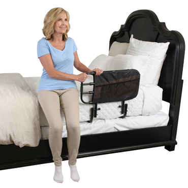 A woman sits on the edge of a bed, holding the Stander EZ Adjust Bed Rail attached to the mattress. The white bedding and bright room highlight her focus on fall protection and bedside safety.