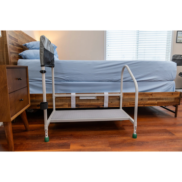 The Step2bed Deluxe Bedside Step-Up Platform with Accessory Kit is placed beside a wooden bed with blue bedding, next to a wooden nightstand, on a wooden floor in a sunlit bedroom.