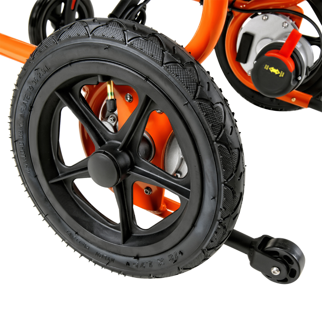 Close-up of a SuperHandy GoRide 2 Portable Electric Wheelchair showing a black treaded tire on an orange frame, reminiscent of a lightweight wheelchair. The wheel has a small black hubcap with a stabilizing training wheel on the right, with engine parts visible in the background.