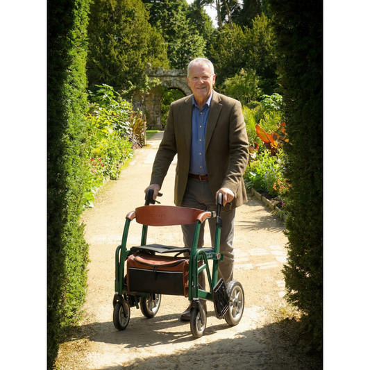 A smiling older man walks along a garden path using a green rollator walker with a padded seat.