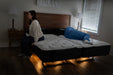 A woman sits pensively on a beds edge in a dimly lit room, while someone rests under a soft Premier mattress atop the Flexabed Hi-Low SL Adjustable Bed. The space features wooden floors and a nightstand with two lamps softly illuminating the scene.