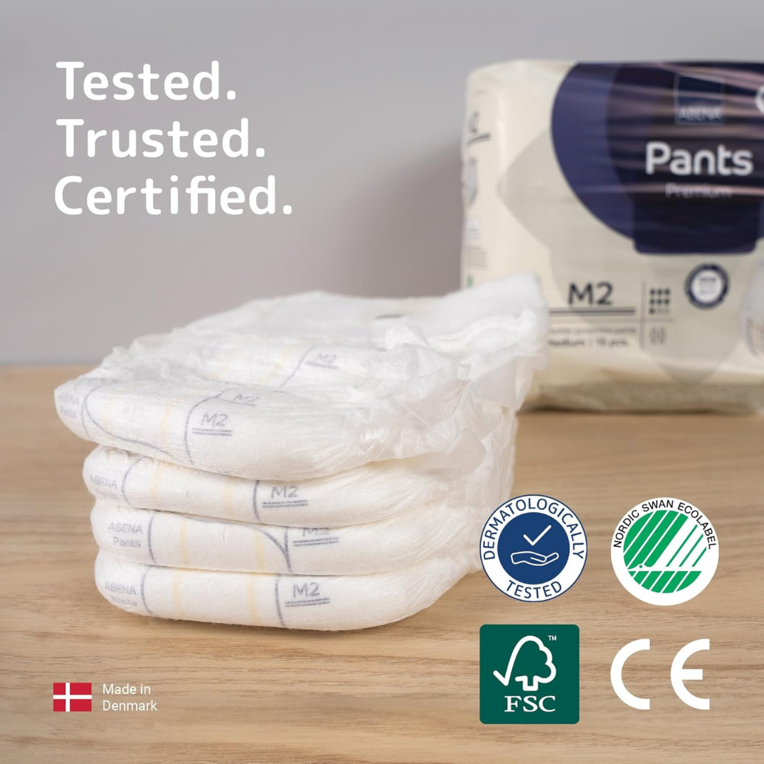 Four folded Abena Pants Premium Protective Adult Underwear sit on a wooden surface before a package labeled "Pants Premium," displaying certification and eco-label logos, with "Tested. Trusted. Certified." and "Made in Denmark" text shown.