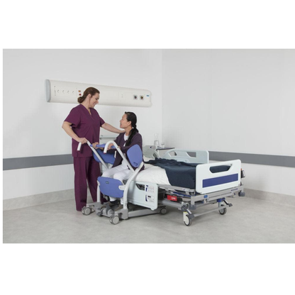 A nurse in purple scrubs assists a patient on the edge of a hospital bed, using the ARJO Sara Stedy Manual Standing Aid & Sit-To-Stand Transfer Lift in a clean, white room, smiling and holding the patient's arm for support.