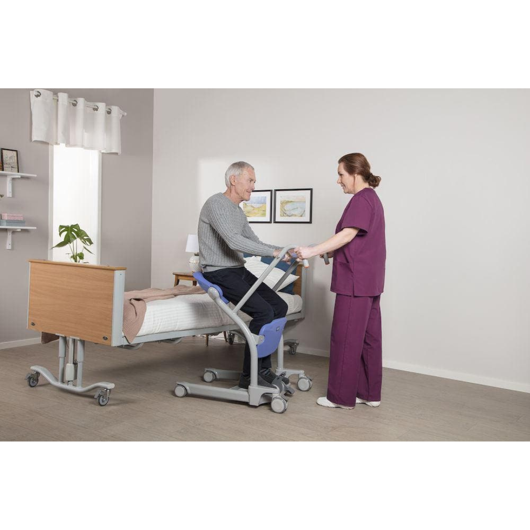A nurse in purple scrubs assists an elderly man using the ARJO Sara Stedy Manual Standing Aid & Sit-To-Stand Transfer Lift next to a hospital bed in a clean, well-lit room with minimal decor.