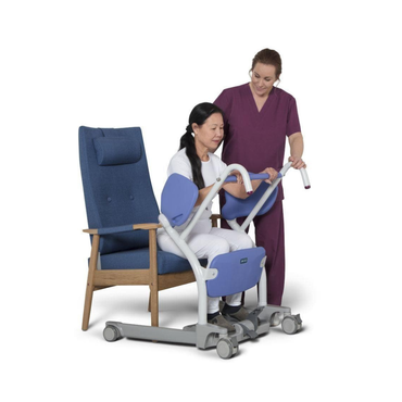 A caregiver in purple scrubs assists a seated woman using the ARJO Sara Stedy Manual Standing Aid & Sit-To-Stand Transfer Lift, positioned in front of a modern blue chair against a white background.