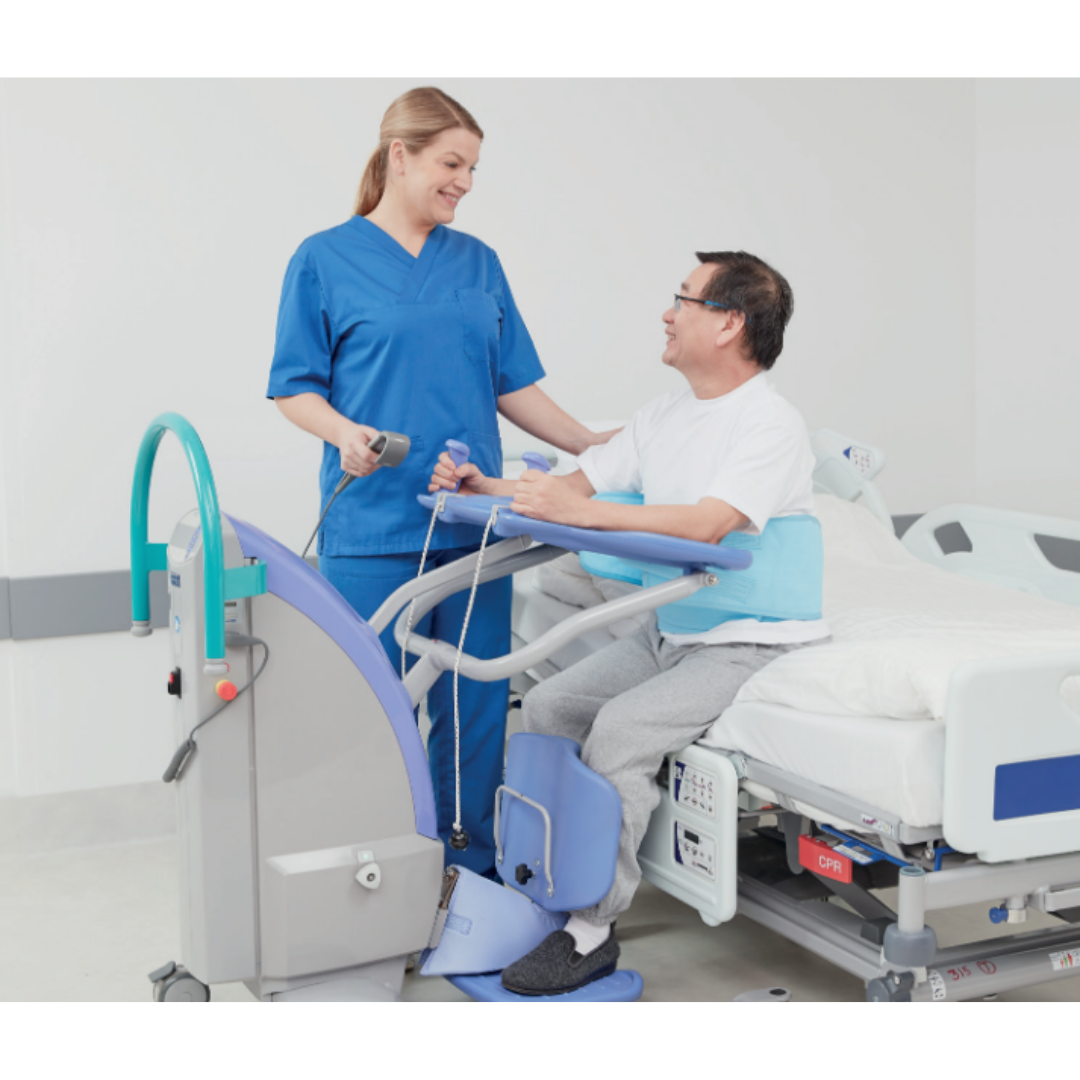A nurse in blue scrubs assists a smiling male patient as he uses the ARJO Sara Plus Powered Sit-To-Stand beside a hospital bed, supporting his mobility and gait training while he holds the device’s handles and partially stands.