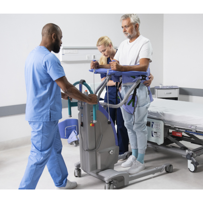 Two healthcare workers assist a man using the ARJO Sara Plus Powered Sit-To-Stand in a hospital room. The device supports him as the workers guide and monitor his mobility for safety. A hospital bed is nearby.