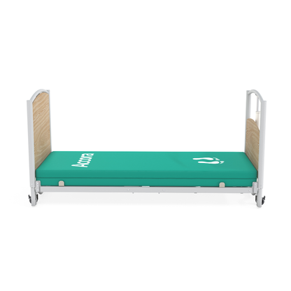 A hospital bed with side rails, light wood headboard and footboard, and a green mattress with white medical symbols and "Antica," styled as an Accora FloorBed® Ultra Low Full Electric Homecare Bed.