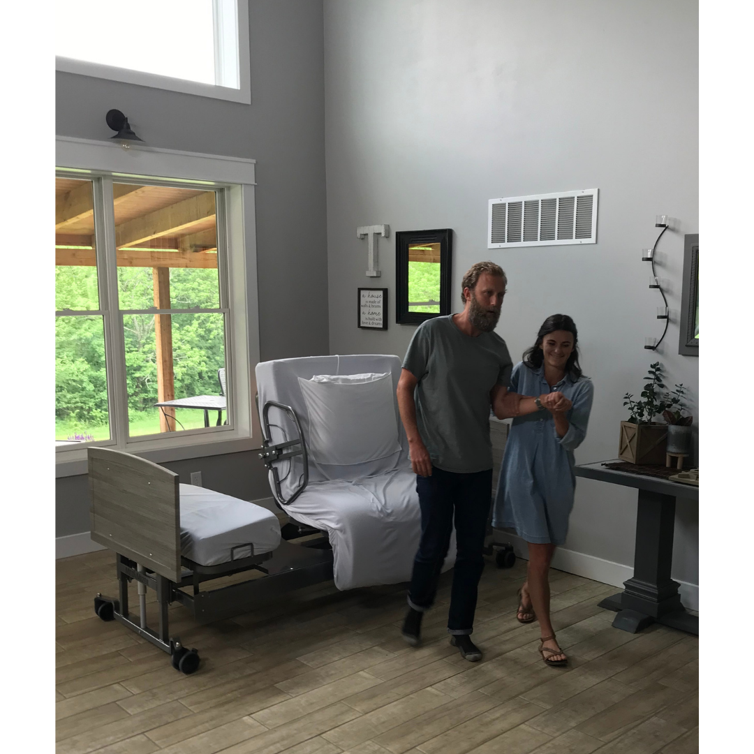 A man supports a woman walking in a living room featuring the Med-Mizer ActiveCare Rotating Hospital Bed by the window. Sunlight highlights wood floors, light walls, and decorations throughout the space.