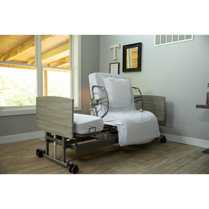 The Med-Mizer ActiveCare Rotating Hospital Bed, featuring light wood panels and white bedding, is placed in a bright, tidy room with large windows and décor—an ideal choice for comfortable home care.