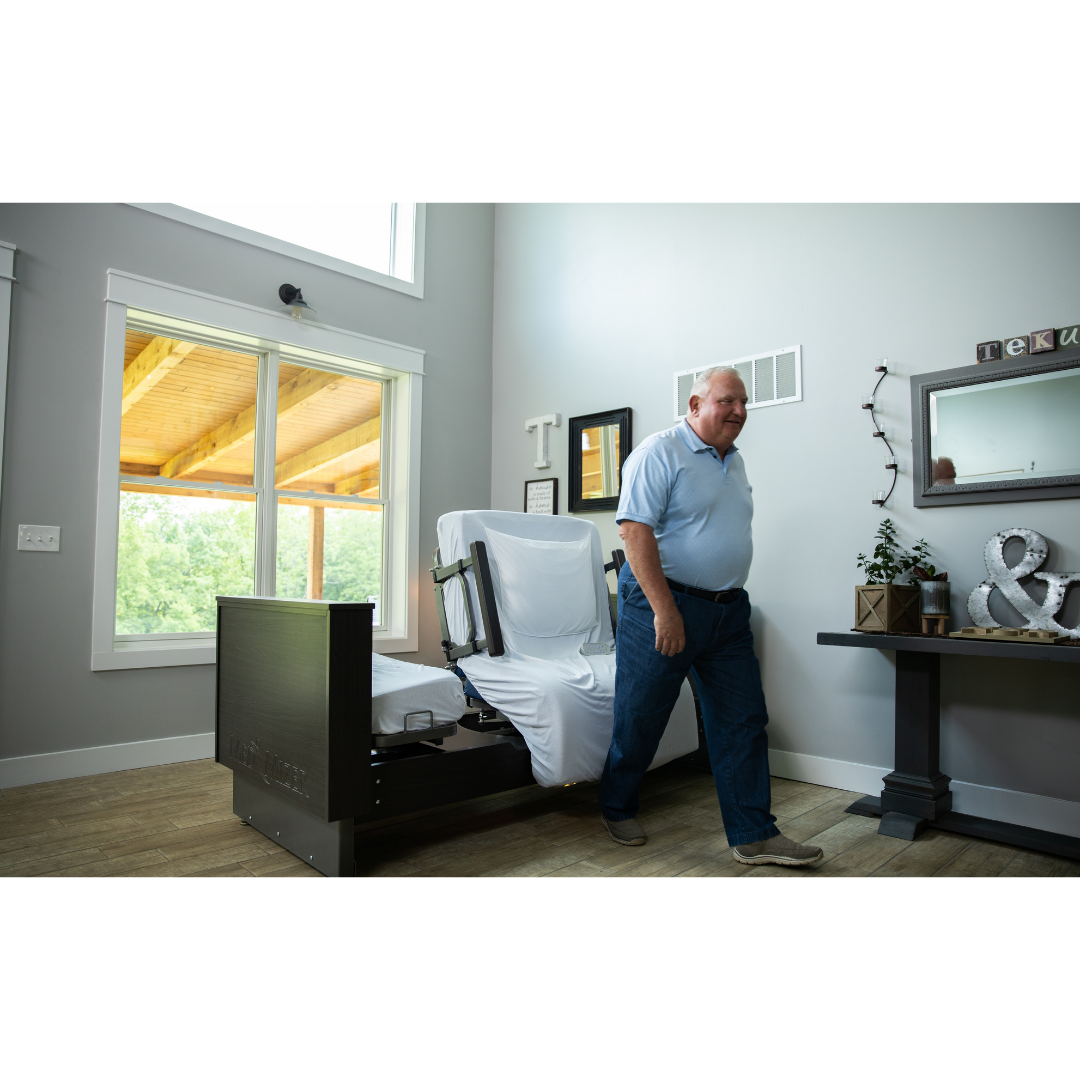 A man in a light blue shirt and jeans walks past a Med-Mizer ActiveCare Rotating Hospital Bed in a bright, modern room with large windows and wooden floors, highlighting comfort beyond the standard hospital bed.