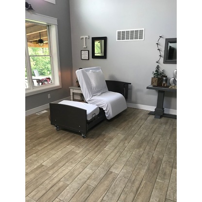 A Med-Mizer ActiveCare Rotating Hospital Bed with white sheets stands in a bright living room featuring wood-style floors, a large window, gray walls adorned with a mirror, artwork, and small plants.