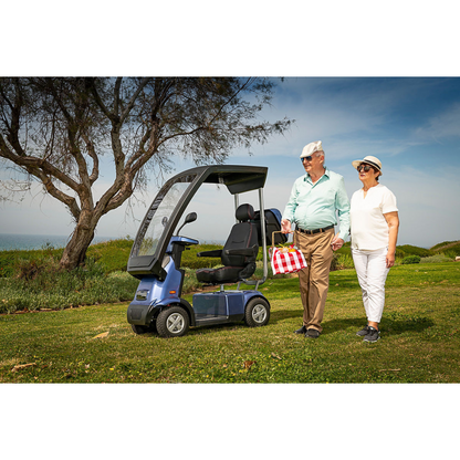 An elderly couple strolls beside the Afikim Afiscooter C4 HD, an electric mobility scooter with a canopy. The man carries a red-checkered bag; both wear hats and sunglasses. An orthopedic seat ensures comfort as they enjoy the scenic view of a tree and blue sky.