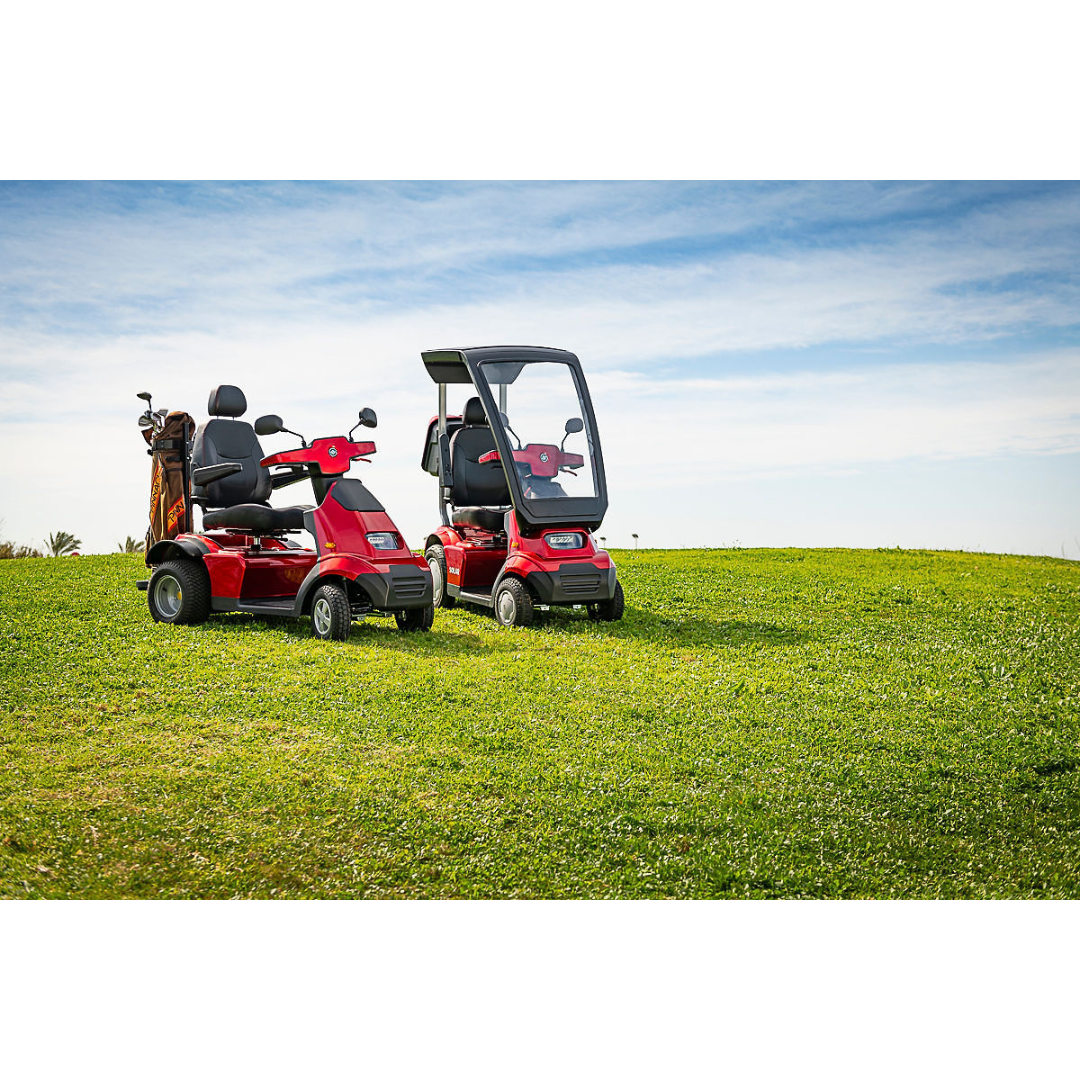 Two red Afikim Afiscooter Solar mobility scooters sit on a grassy hill under a blue sky. The right one features a transparent windshield, roof, and orthopedic seat for added comfort, while the left is open. Both are positioned side by side, facing slightly to the right.