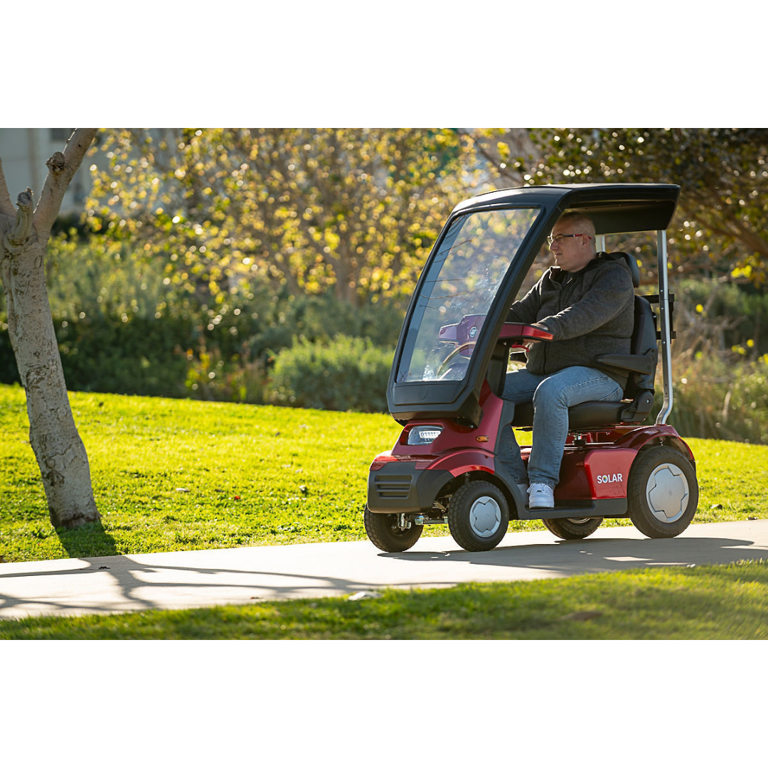 A person navigates a grassy park in a small red Afikim Afiscooter Solar, a solar-powered bariatric mobility scooter with a canopy. The transparent windshield glistens under the suns rays, harnessed for sustainable energy, casting playful shadows on the ground.