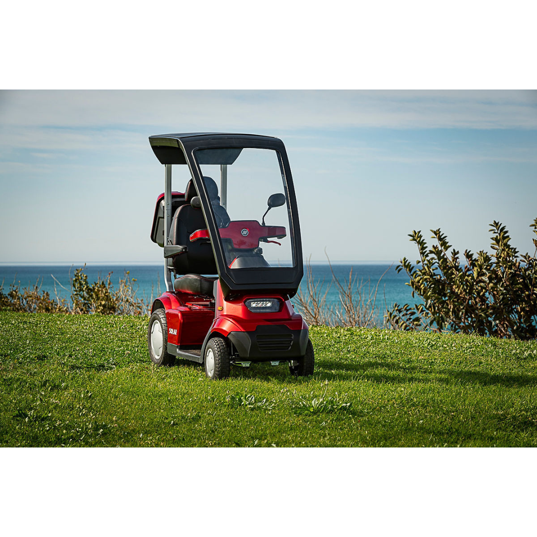 The Afikim Afiscooter Solar, a red solar-powered bariatric mobility scooter with a canopy, is parked on grass. The scene includes an ocean view under a blue sky with clouds and shrubs on the right.