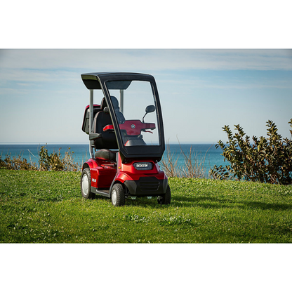 The Afikim Afiscooter Solar, a red solar-powered bariatric mobility scooter with a canopy, is parked on grass. The scene includes an ocean view under a blue sky with clouds and shrubs on the right.