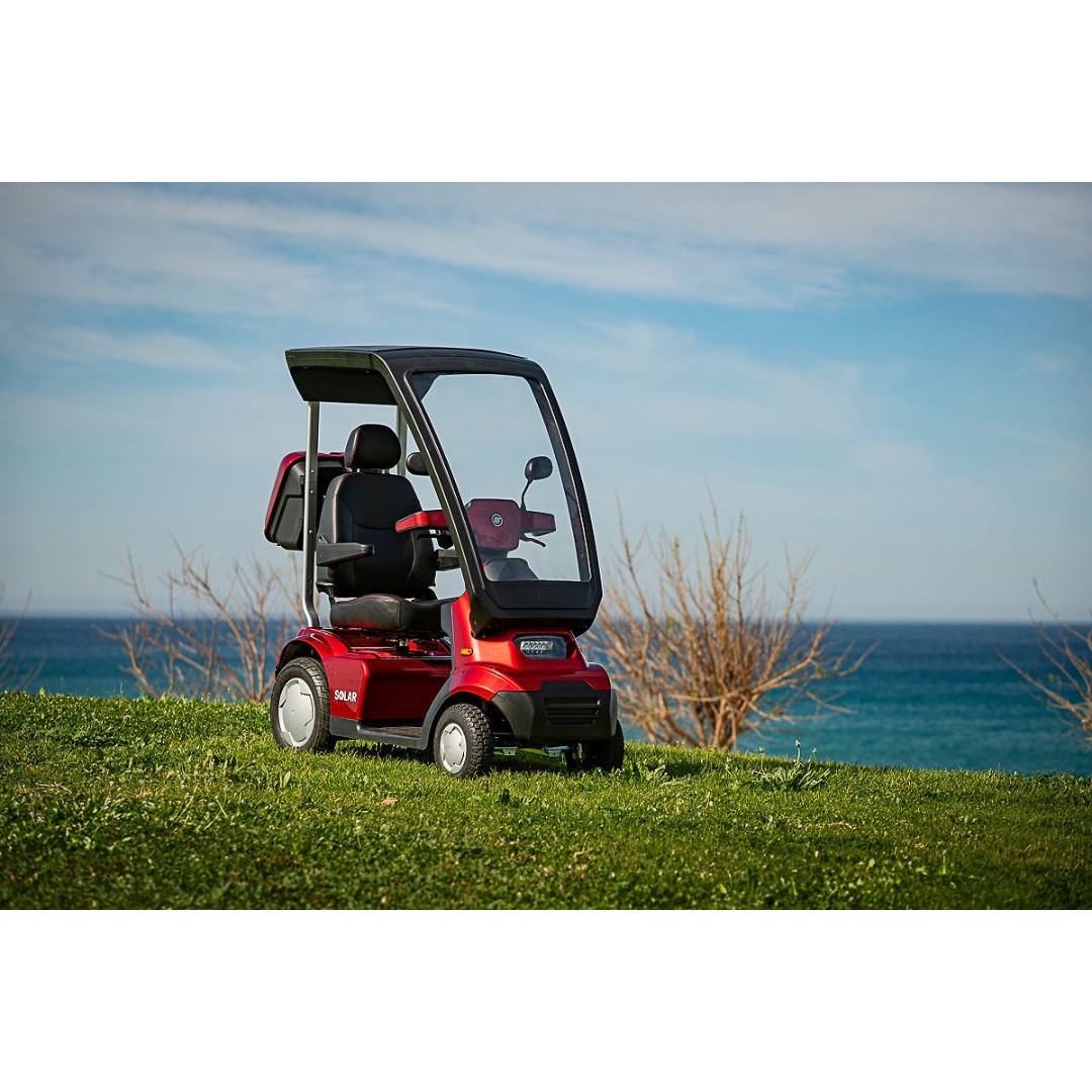 An Afikim Afiscooter Solar, a solar-powered bariatric mobility scooter with a transparent canopy, is parked on a grassy hillside overlooking the serene sea. The partly cloudy sky and calm ocean add to the tranquil scene, with bare shrubs visible in the distance.