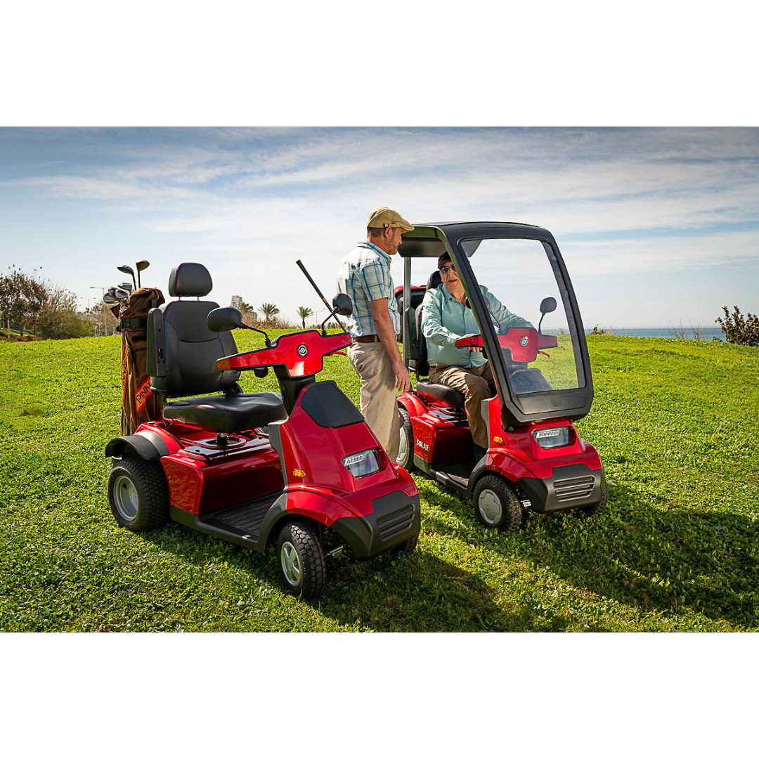 Two people are on Afikim Afiscooter Solar mobility scooters, highlighting solar charging efficiency, on a grassy area. One scooter has a clear canopy; the other is open and carries golf clubs. They are side by side, with the blue sky and ocean as a stunning backdrop.