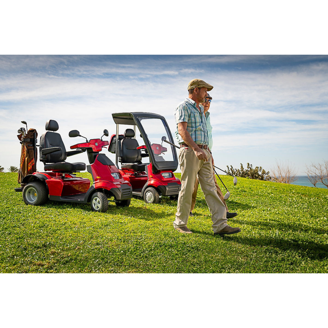 Two people stroll on a golf course; one carries a club. Nearby, two Afikim Afiscooter Solar mobility scooters, one featuring a canopy, are parked on the lush green grass. A clear sky reveals the ocean in the distance.